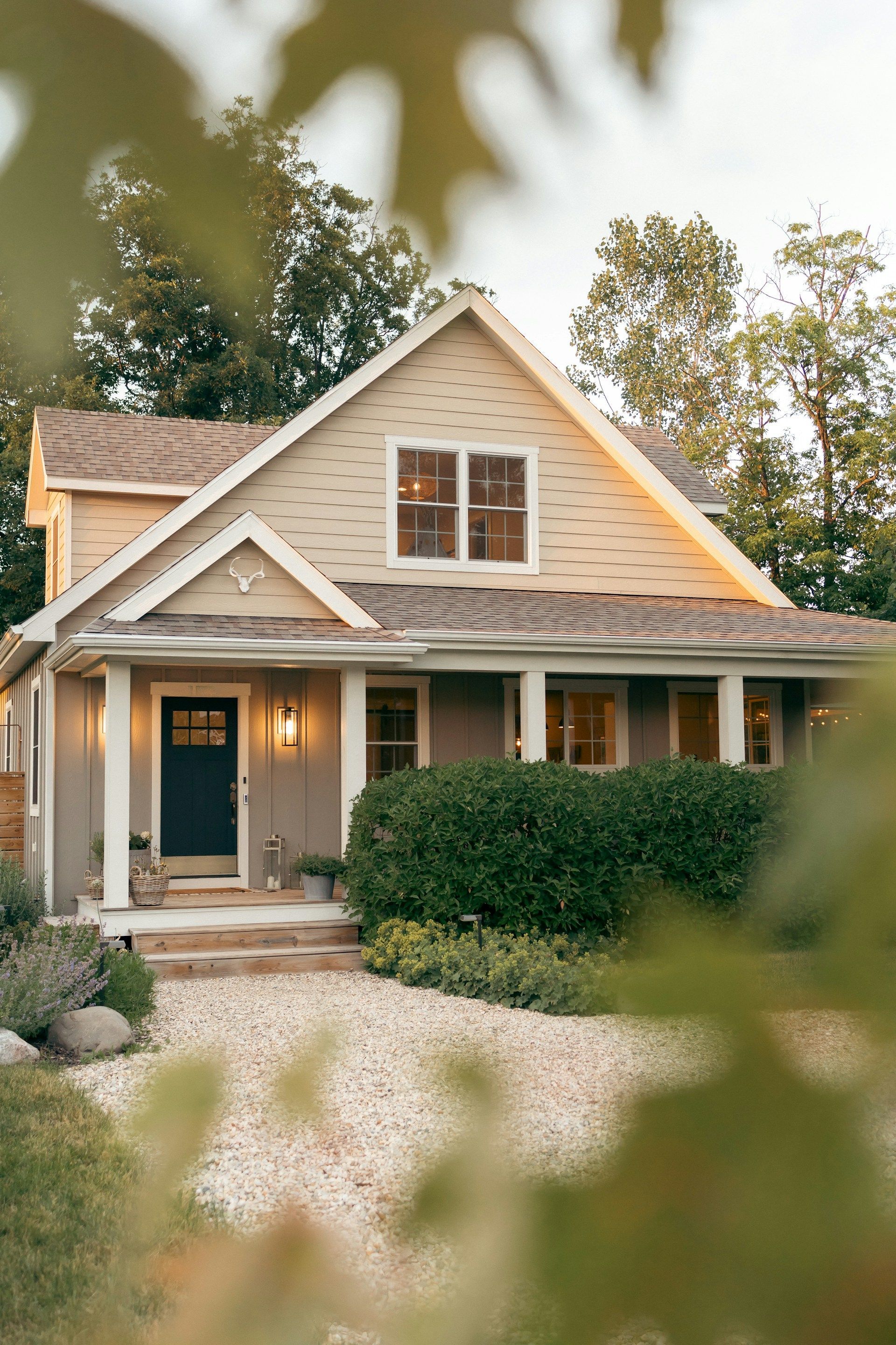 Cozy, light-colored house with a porch. A dark blue door is centered under a peaked roof with windows. Bushes and gravel surround.
