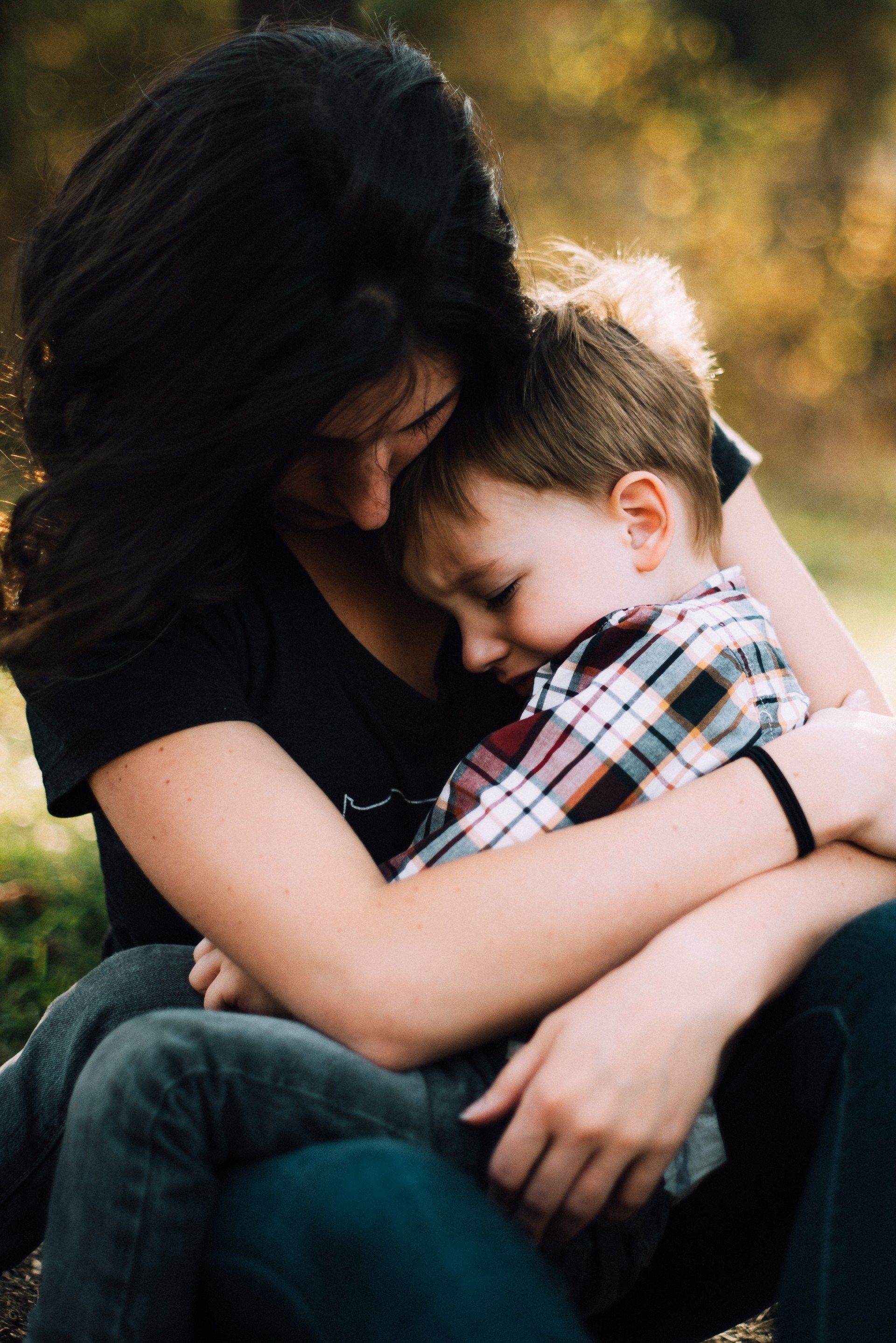 Woman embracing a young child, both seated outdoors. The child is sad, leaning on the woman's shoulder.