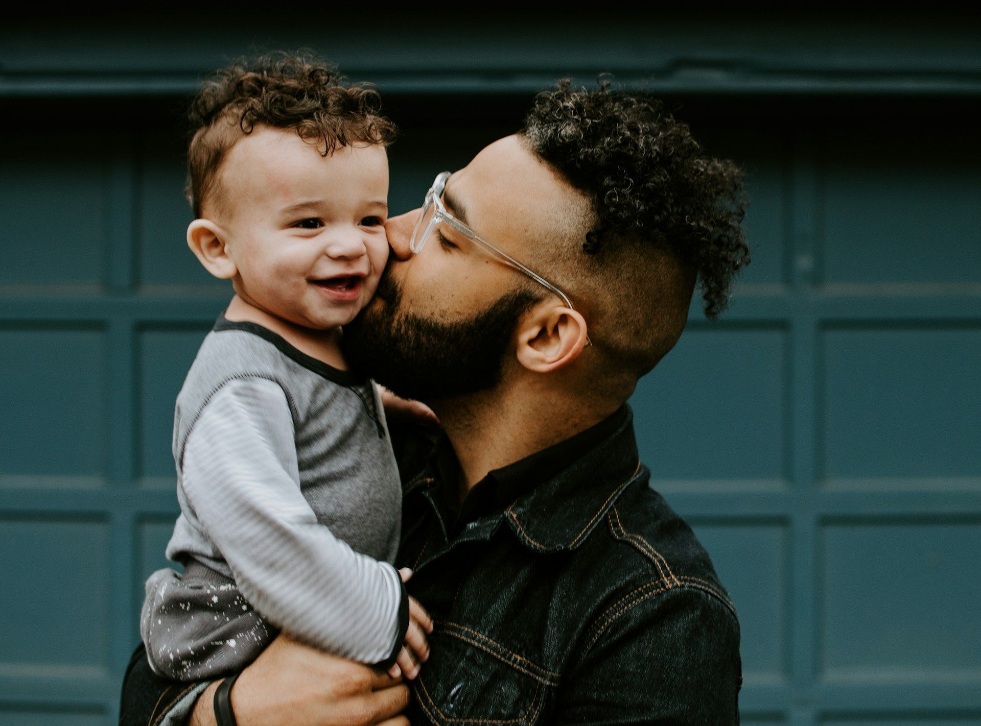Father kisses his smiling young child on the cheek. They stand in front of a blue garage door.