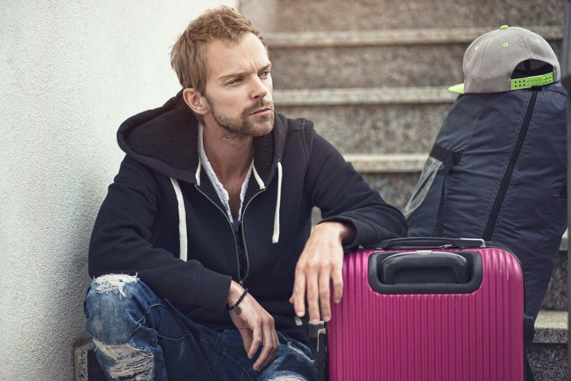 Man in ripped jeans sits on steps with suitcase and backpack, looking thoughtful.