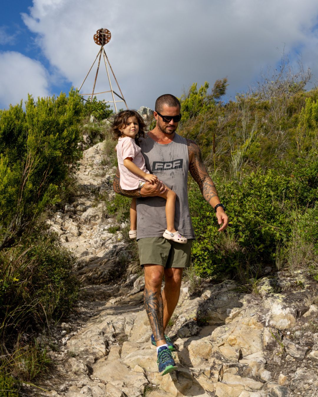 Man carrying a child uphill on a rocky trail with greenery, under a cloudy sky.