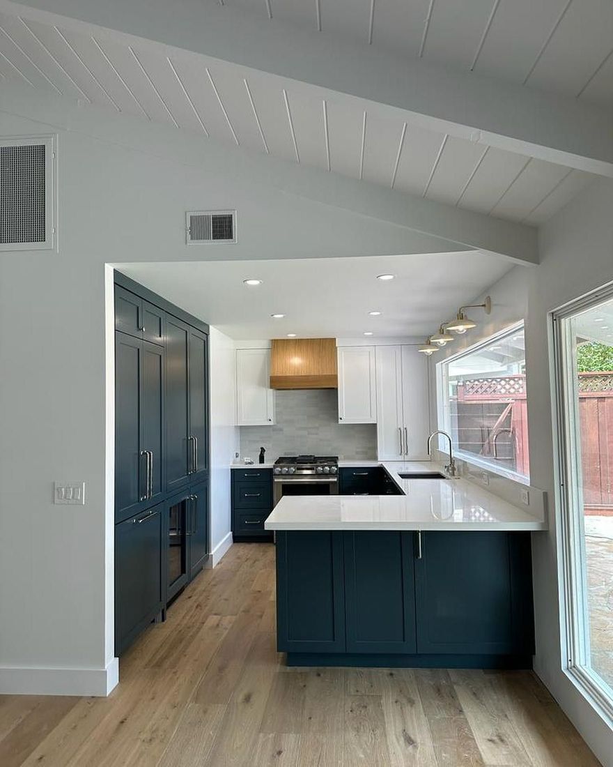 A modern kitchen features dark navy cabinets, white countertops, a wooden range hood, and light wood-tone flooring.