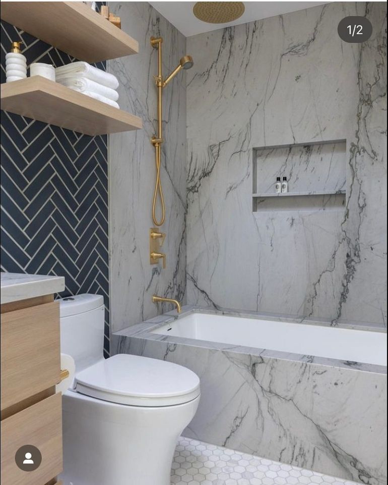 Modern bathroom featuring a marble-tiled bathtub, gold fixtures, dark herringbone accent wall, and light wood shelving.