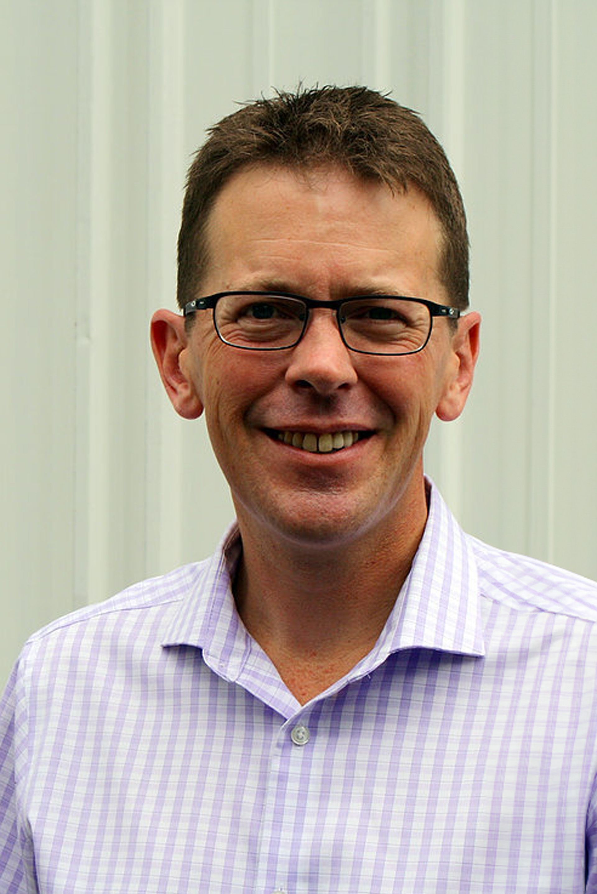 Man with glasses smiles, wearing a light purple plaid shirt, standing in front of a white wall.