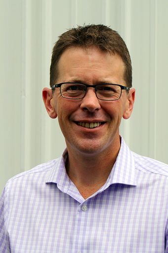 Man with glasses smiles, wearing a light purple plaid shirt, standing in front of a white wall.