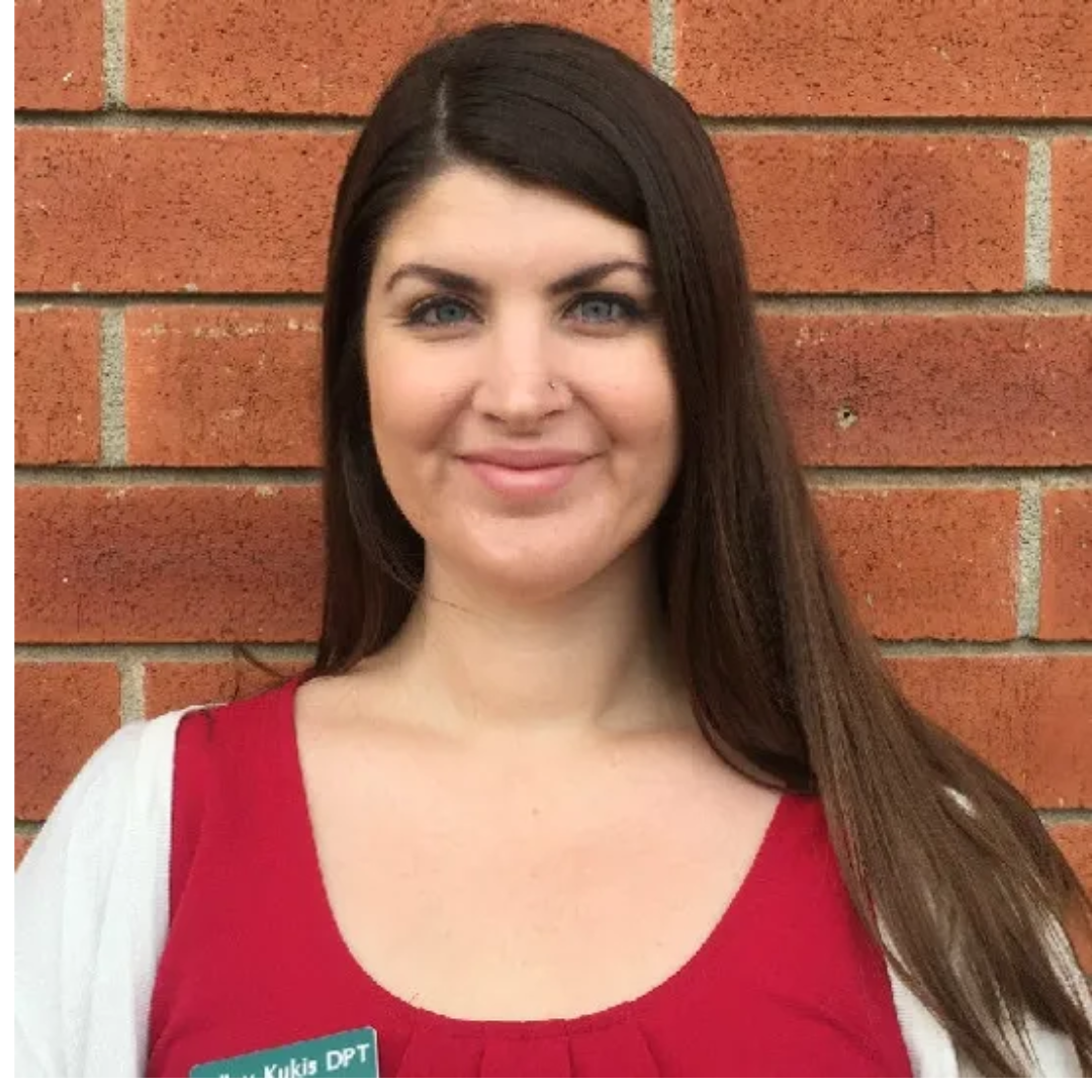 A woman in a red shirt is smiling in front of a brick wall