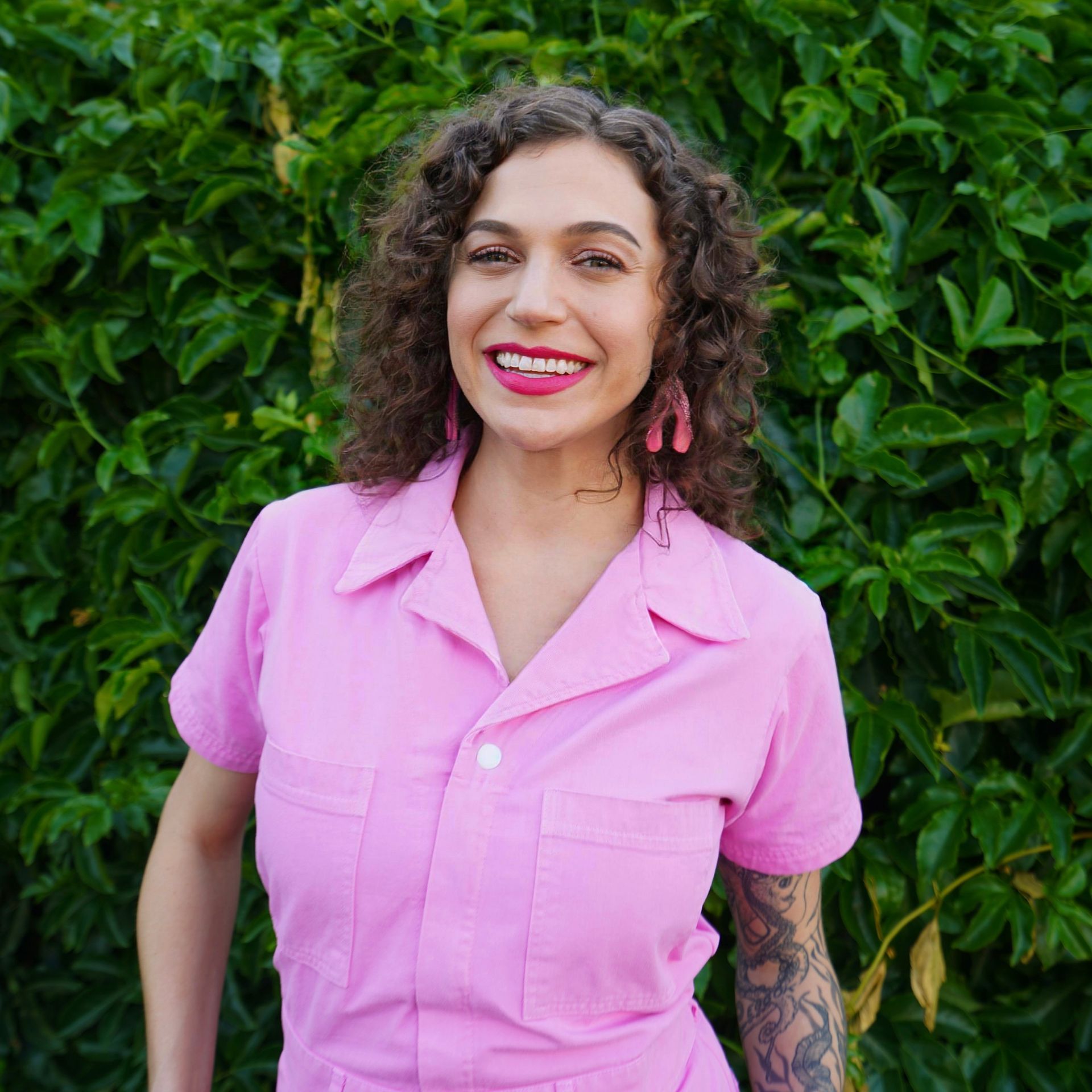 A woman in a pink shirt is smiling in front of a bush.