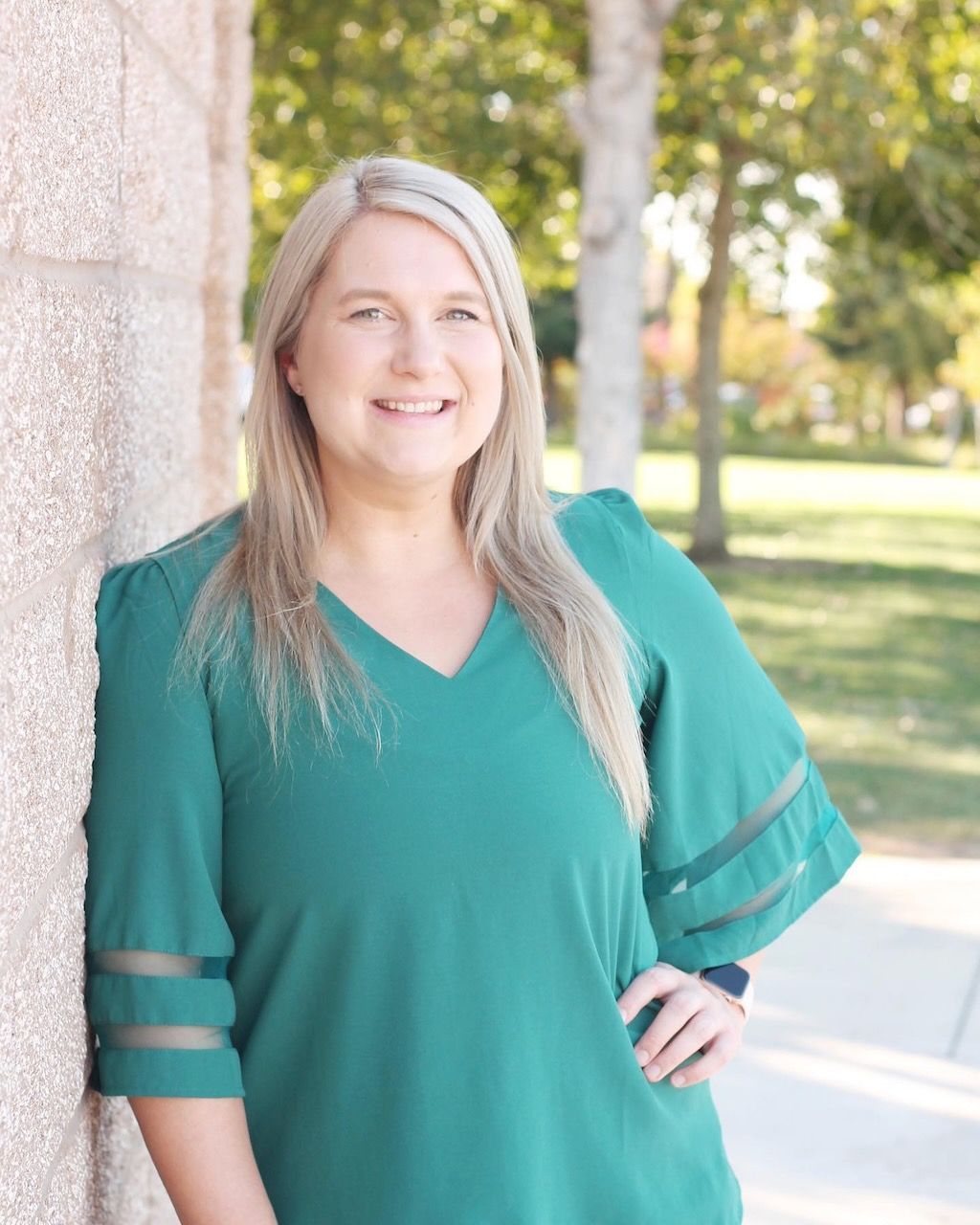 A woman in a green shirt is leaning against a wall and smiling.