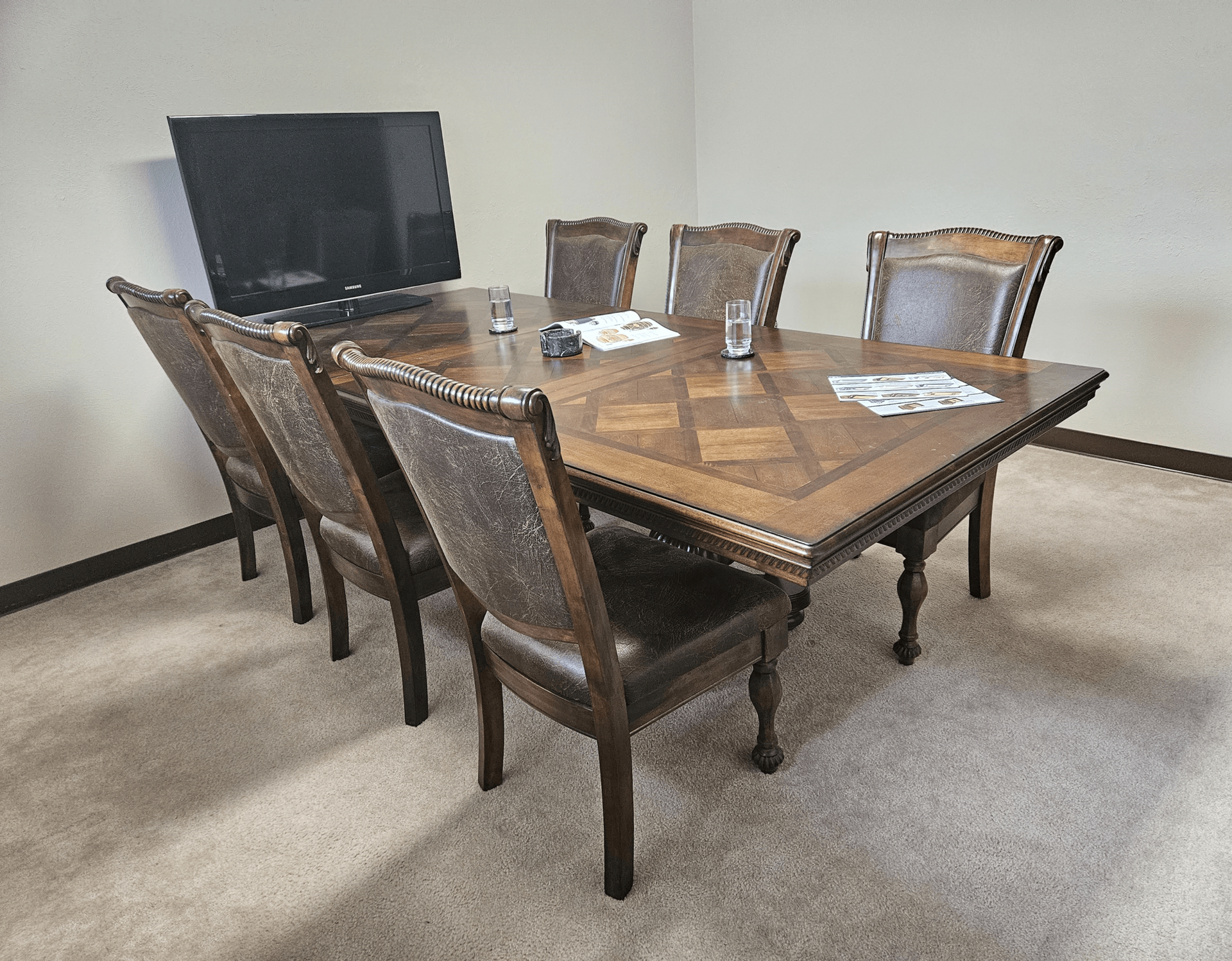 Room with an ornate wooden table and eight upholstered chairs. A TV is against the wall.
