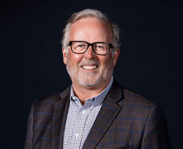 A professional headshot of a smiling person wearing glasses, a patterned button-up shirt, and a dark plaid blazer.