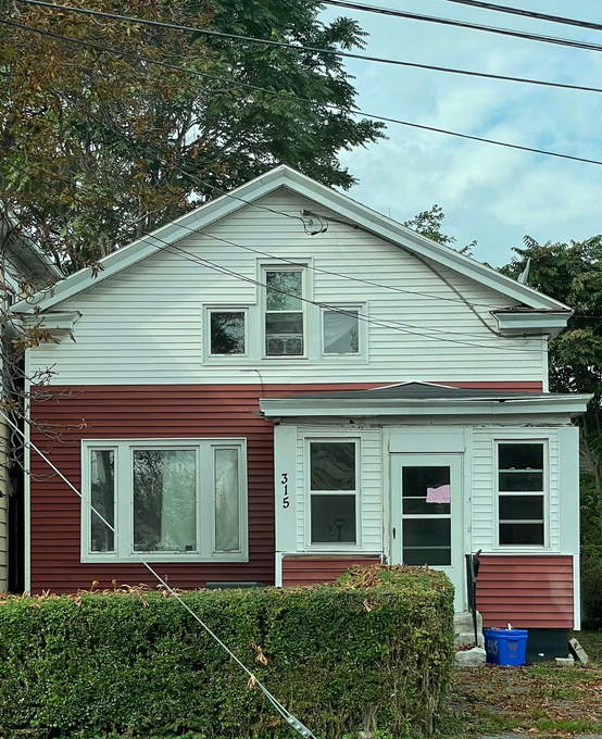 A two-story house with white and dark red siding, a front porch, and a hedge in the front yard.