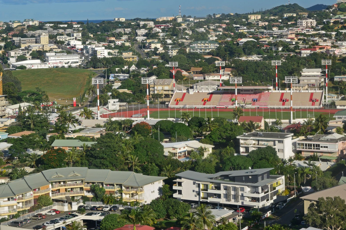 An aerial view of a tropical city featuring a stadium with red seats surrounded by residential buildings and lush trees.
