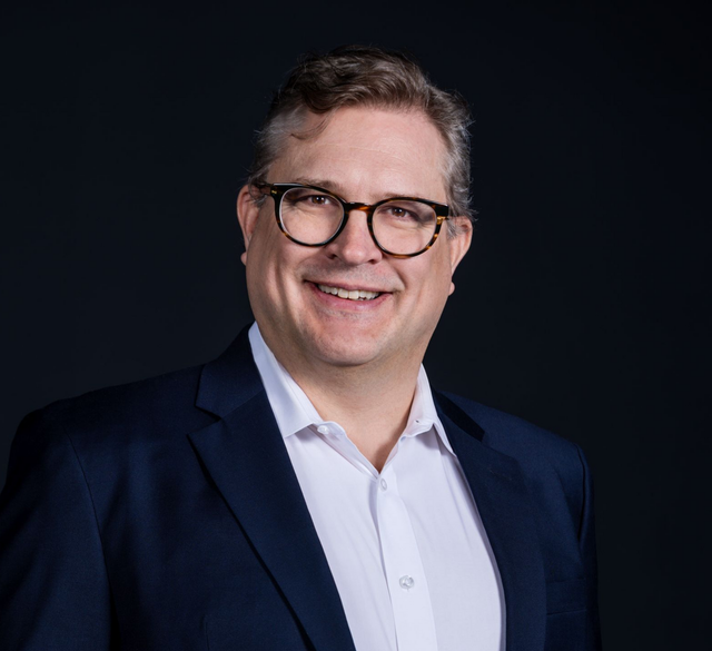 A professional headshot of a person wearing glasses and a dark blue blazer over a white dress shirt, smiling at camera.