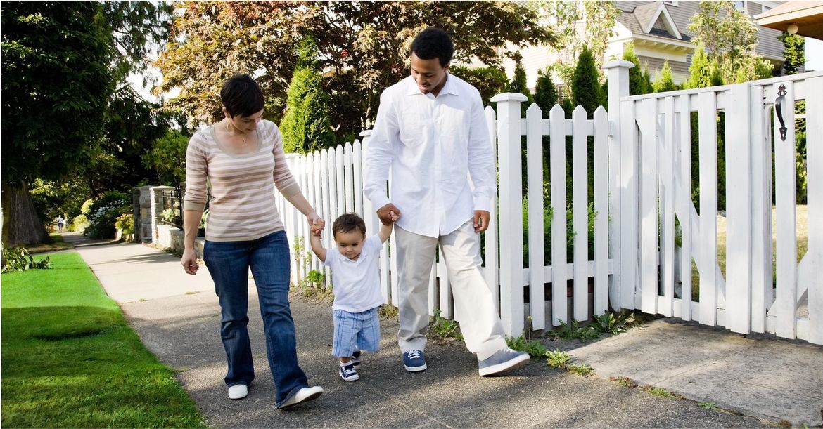 A family of three walks together on a sidewalk past a white picket fence.