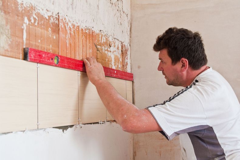 A person uses a red spirit level to check the alignment of beige tiles on a wall during a home renovation.