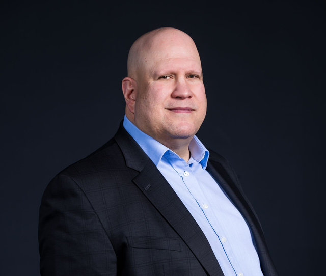 A person with a shaved head wearing a light blue dress shirt and a dark blazer against a plain dark background.