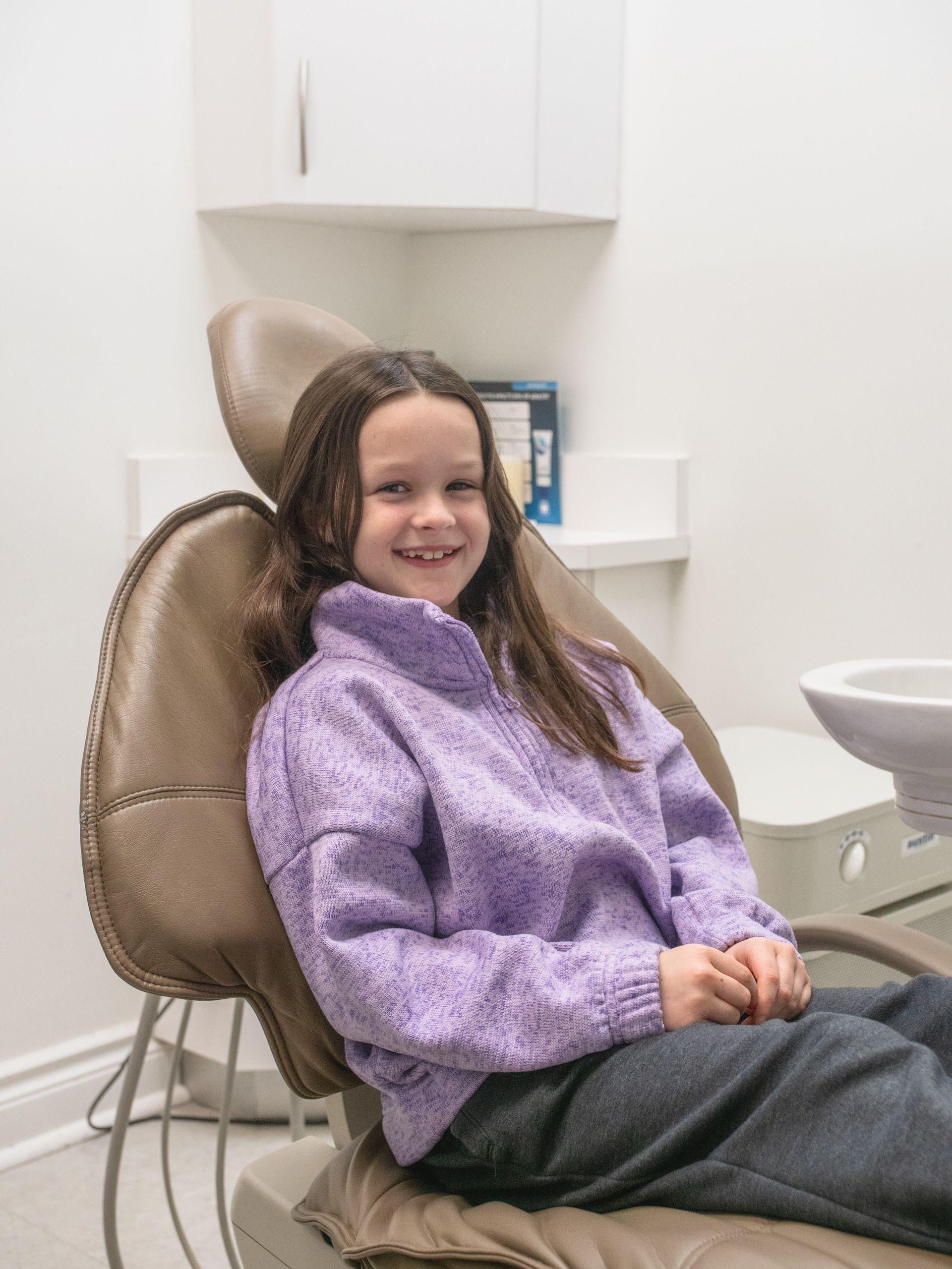 A young girl in a purple sweater is sitting in a dental chair.