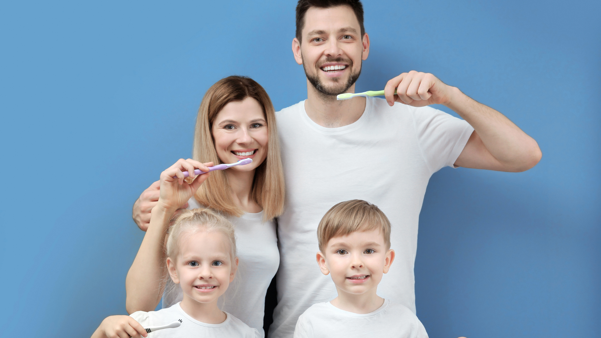 Family of four brushing teeth in front of a blue wall, smiling.