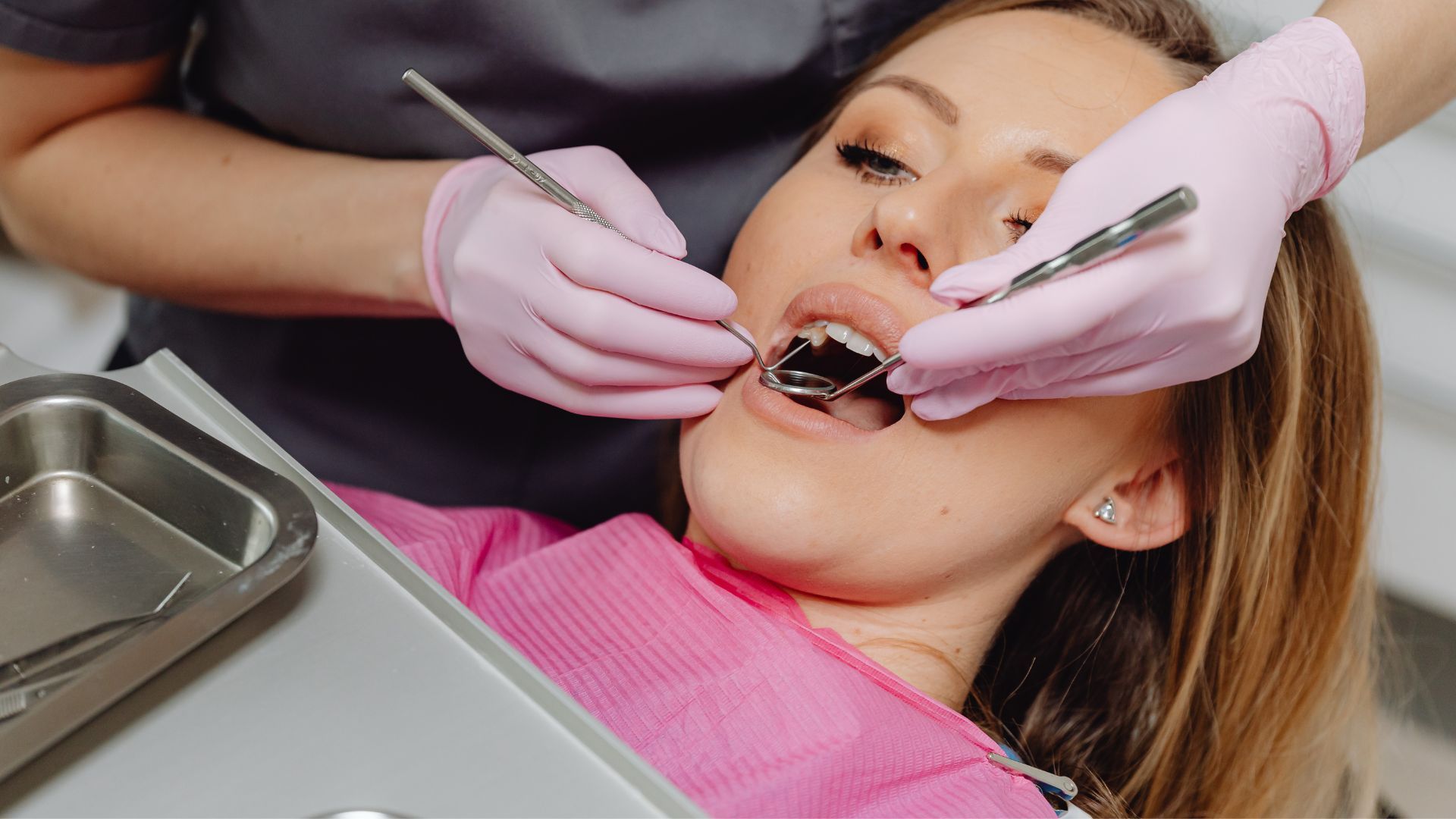 Dentist examining a patient’s mouth with dental tools, pink gloves, and a metal tray in view.