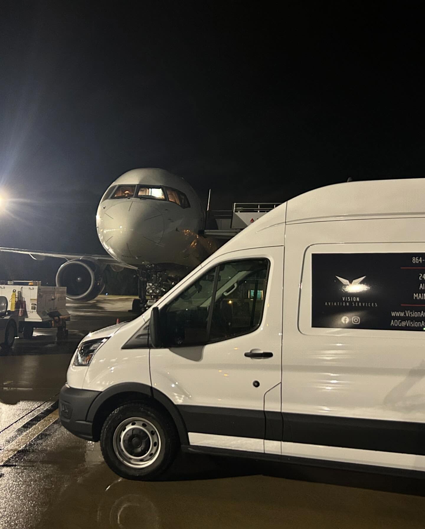 A white van is parked in front of an airplane at night