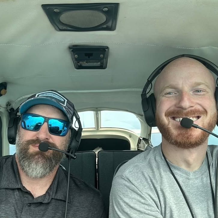 Two men wearing headphones and sunglasses are sitting in a plane.