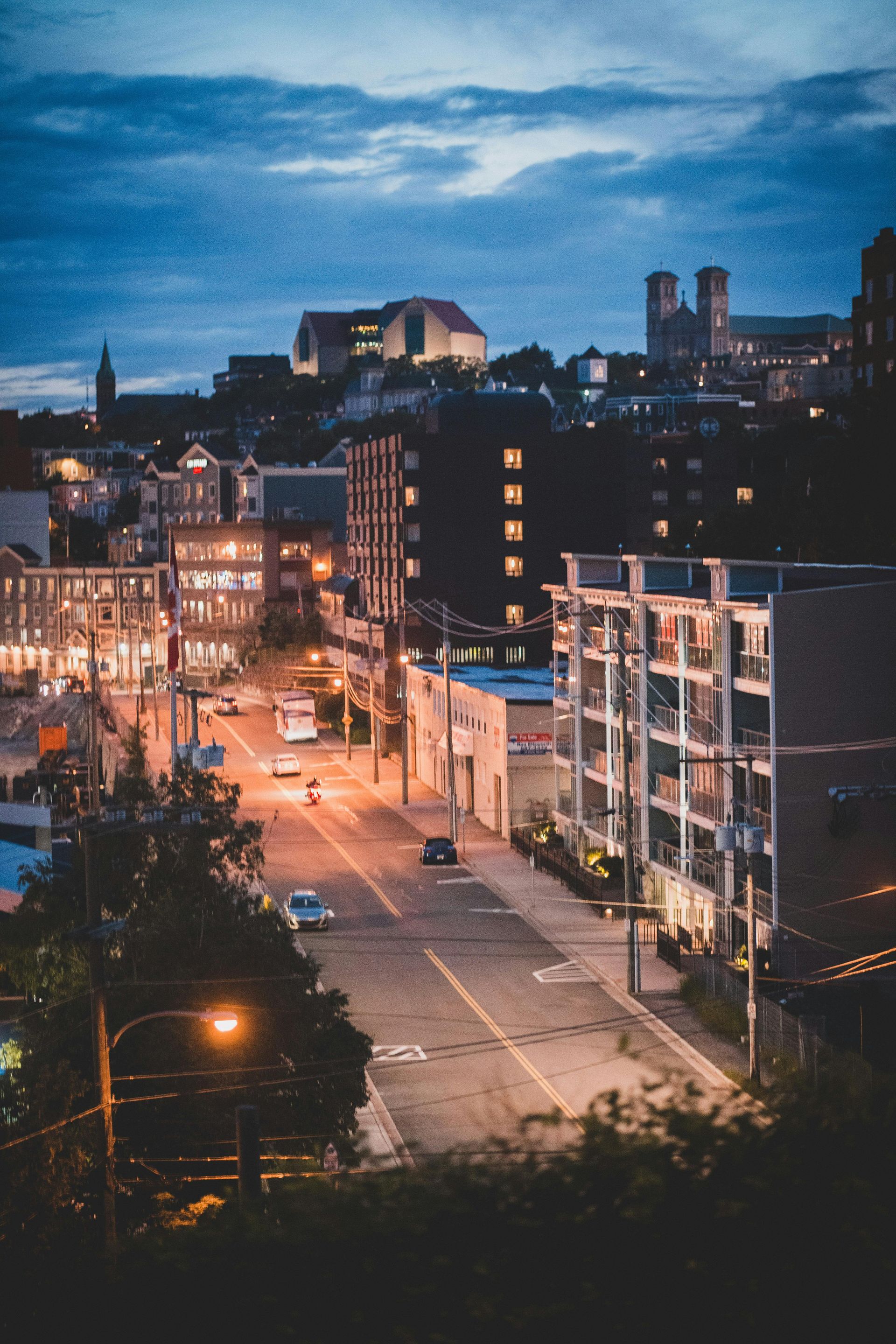 A street scene at twilight with warm streetlights illuminating a road lined with buildings and a town on a distant hill.