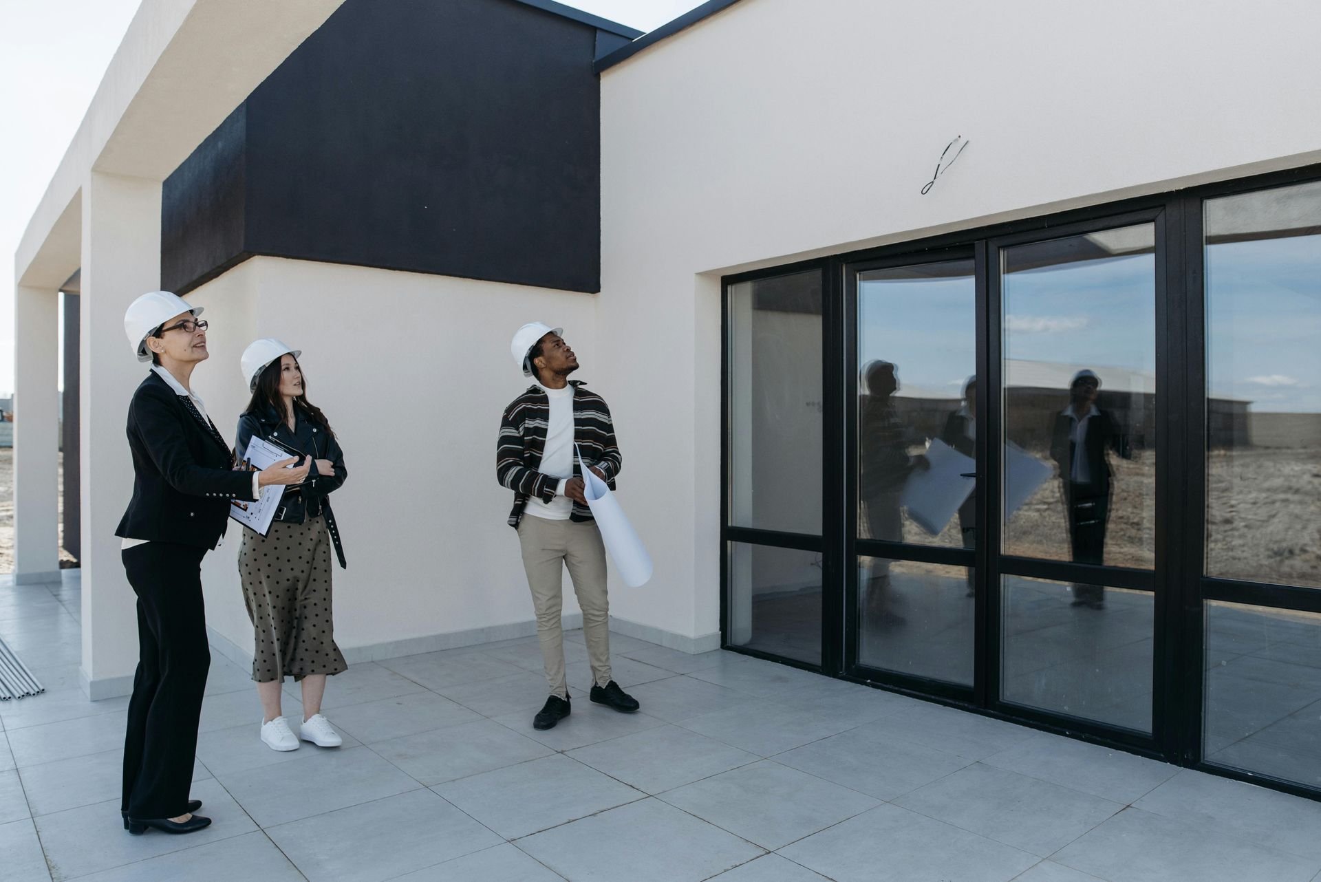 Three people in hard hats stand on a patio, looking at the exterior of a modern building with large glass doors.