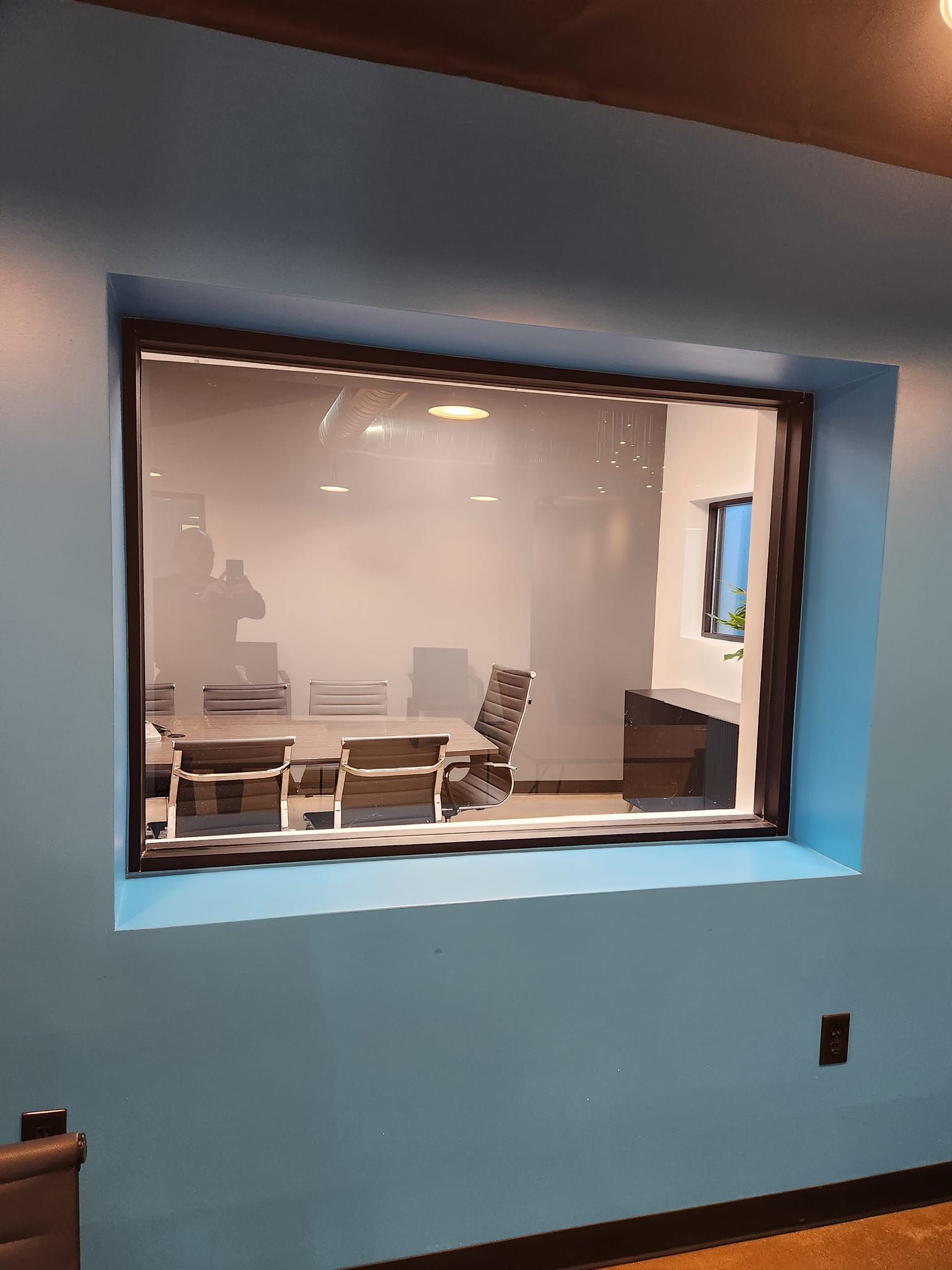 A large window in a blue wall shows a conference room with tables and chairs.