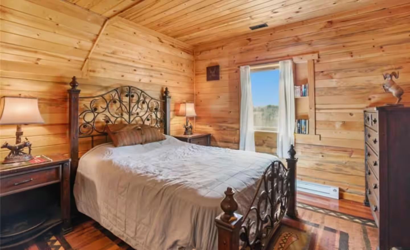A bedroom in a log cabin with a bed , nightstand , dresser and window.