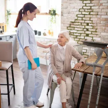 A nurse is talking to an elderly woman who is sitting at a table with crutches.