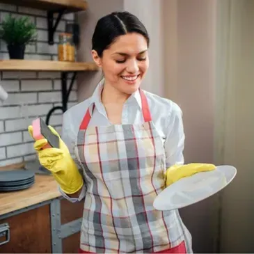 A woman in an apron and yellow gloves is holding a plate and a sponge.