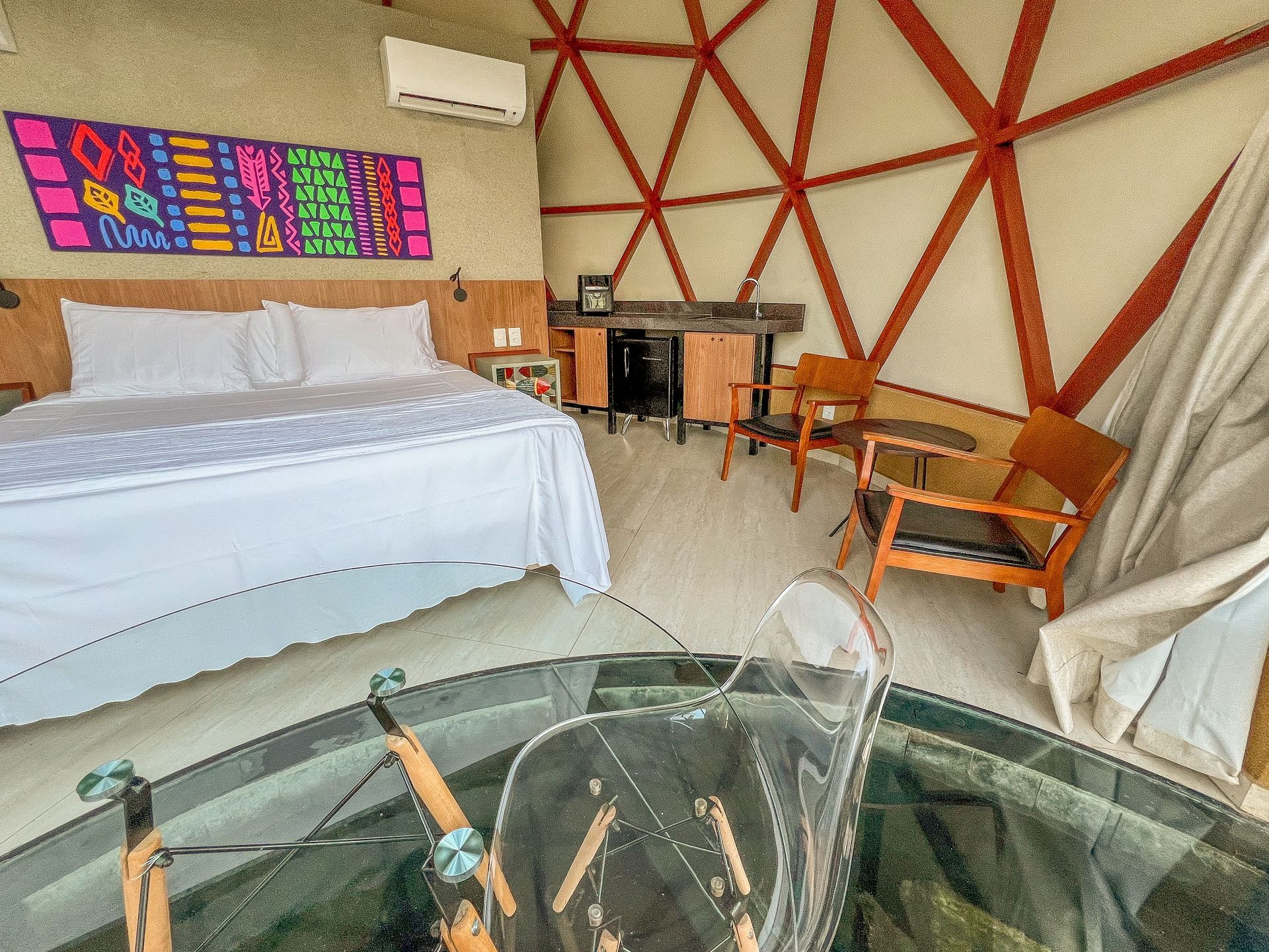 Quarto moderno com cama branca, obras de arte coloridas na parede, cadeiras de madeira e banheira de imersão com box de vidro.