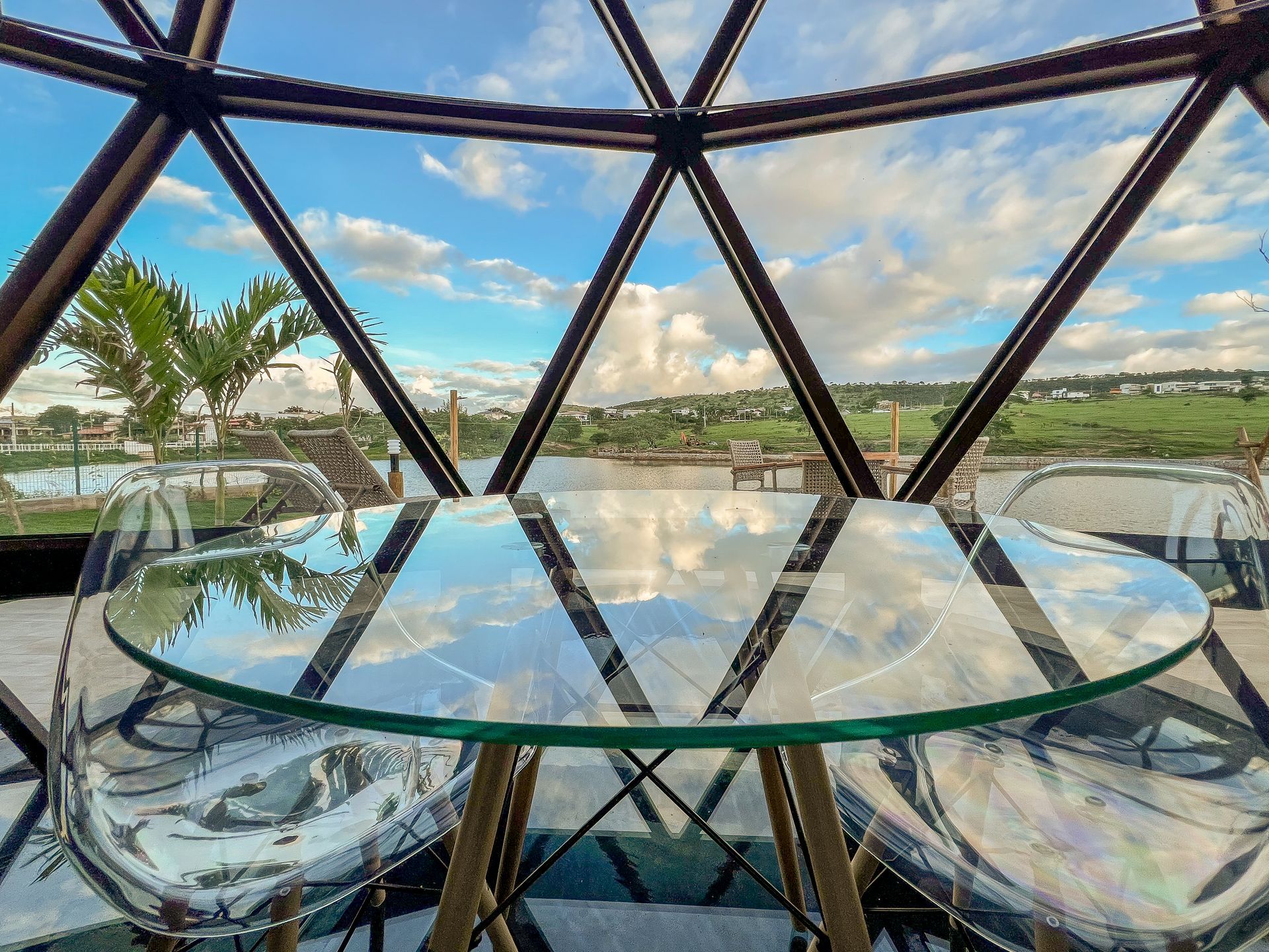Mesa de jantar com cúpula de vidro e duas cadeiras transparentes com vista para um campo de golfe ajardinado e céu azul.