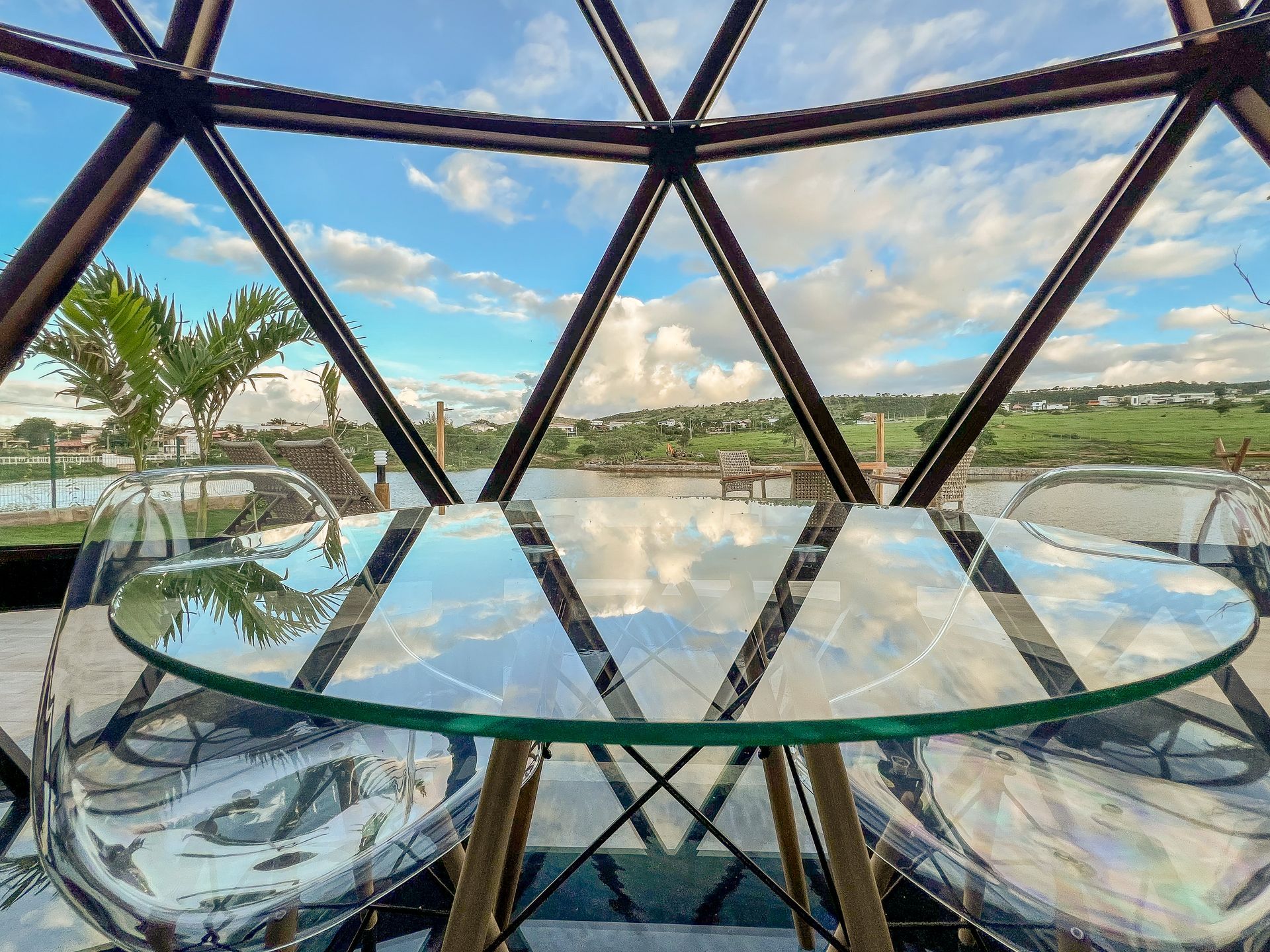 Mesa e cadeiras de vidro dentro de uma cúpula geodésica, com vista para uma piscina ensolarada e paisagem.