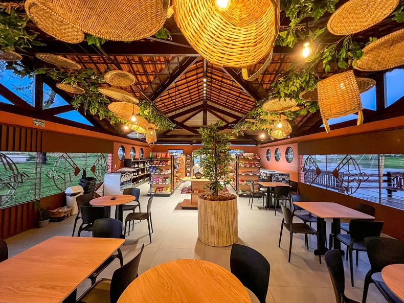 Café ao ar livre com vigas de madeira, luminárias de teto trançadas e mesas centrais com exposição de lanches e bebidas.