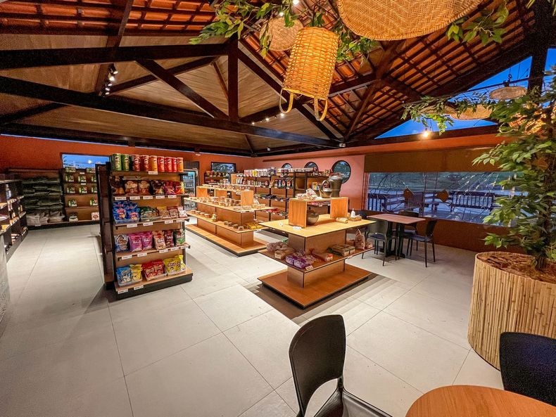 Café ao ar livre com vigas de madeira, luminárias de teto trançadas e mesas centrais com exposição de lanches e bebidas.