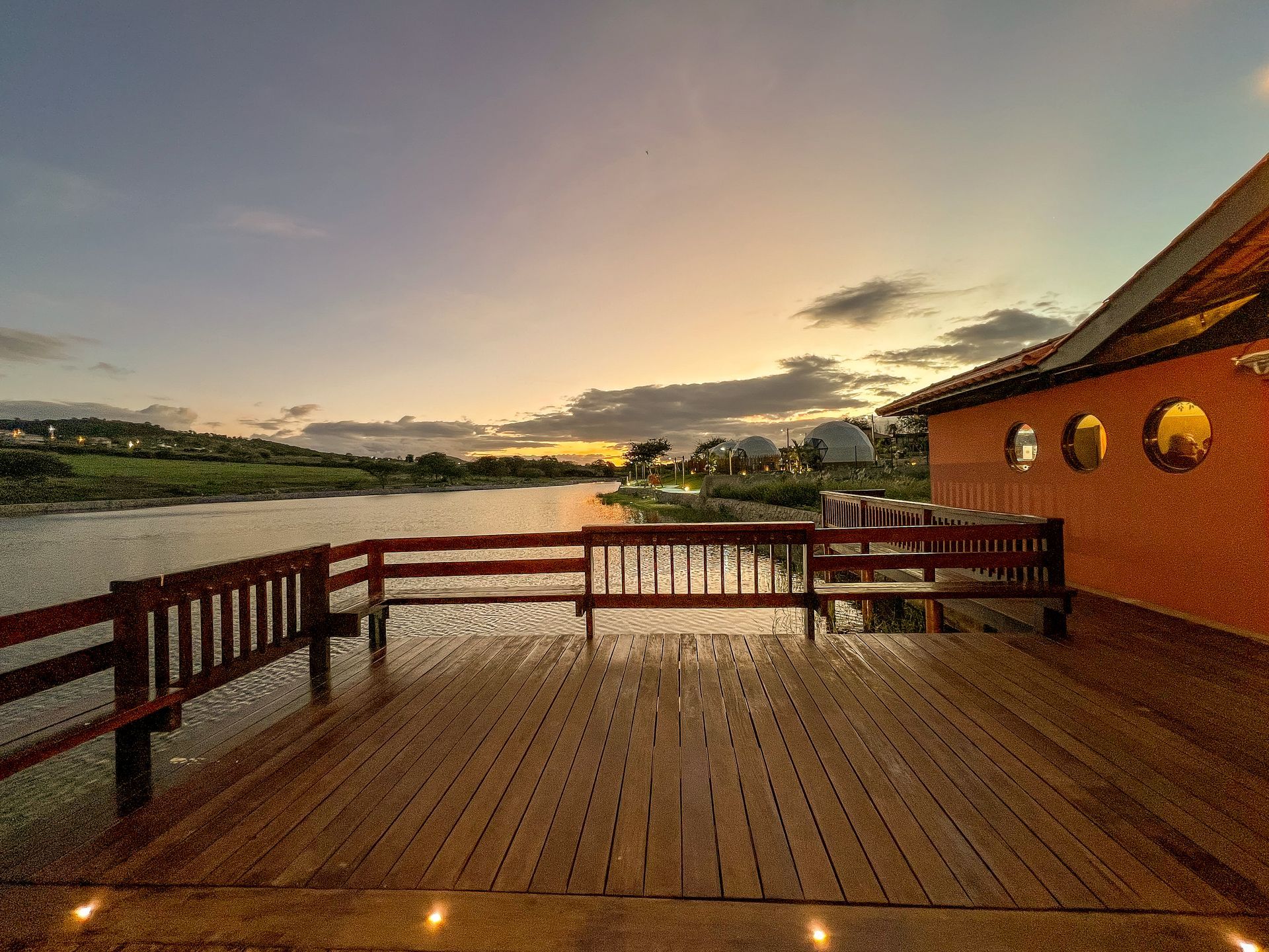 Deck de madeira ao lado de um lago tranquilo ao pôr do sol, com um corrimão vermelho e um prédio de tijolos com janelas redondas.
