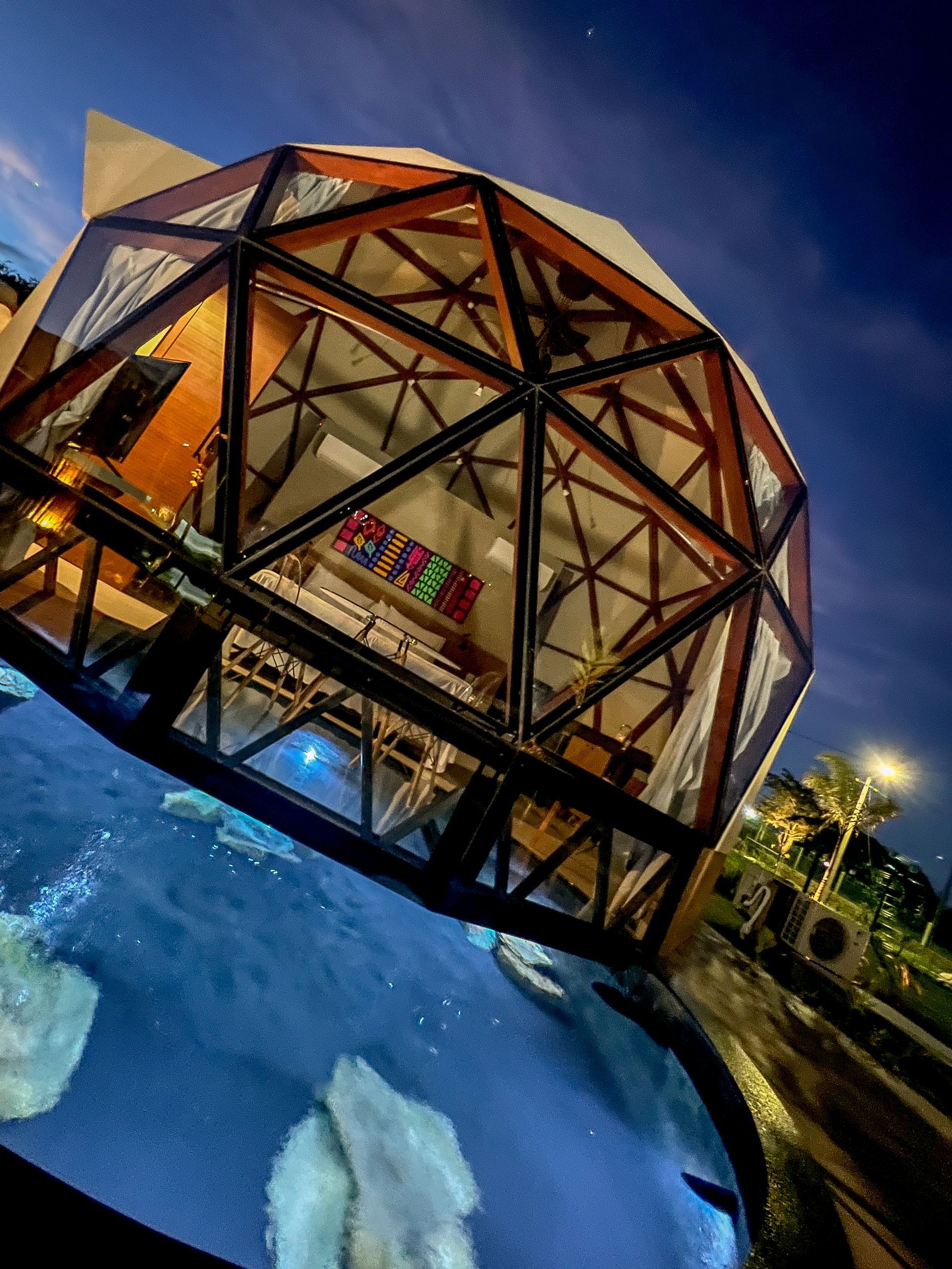 Edifício em forma de cúpula geodésica iluminado ao entardecer, ao lado de uma piscina azul, com árvores e céu noturno.