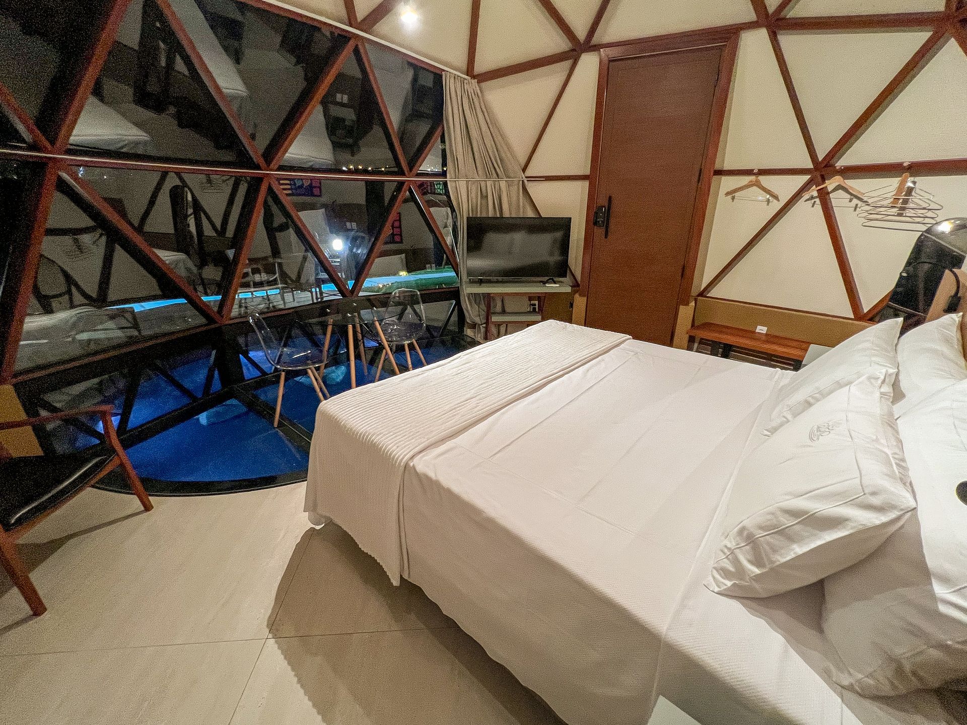 Quarto moderno com cama branca, paredes de madeira com padrões geométricos e janelas do chão ao teto com vista para um ambiente iluminado em tons de azul.