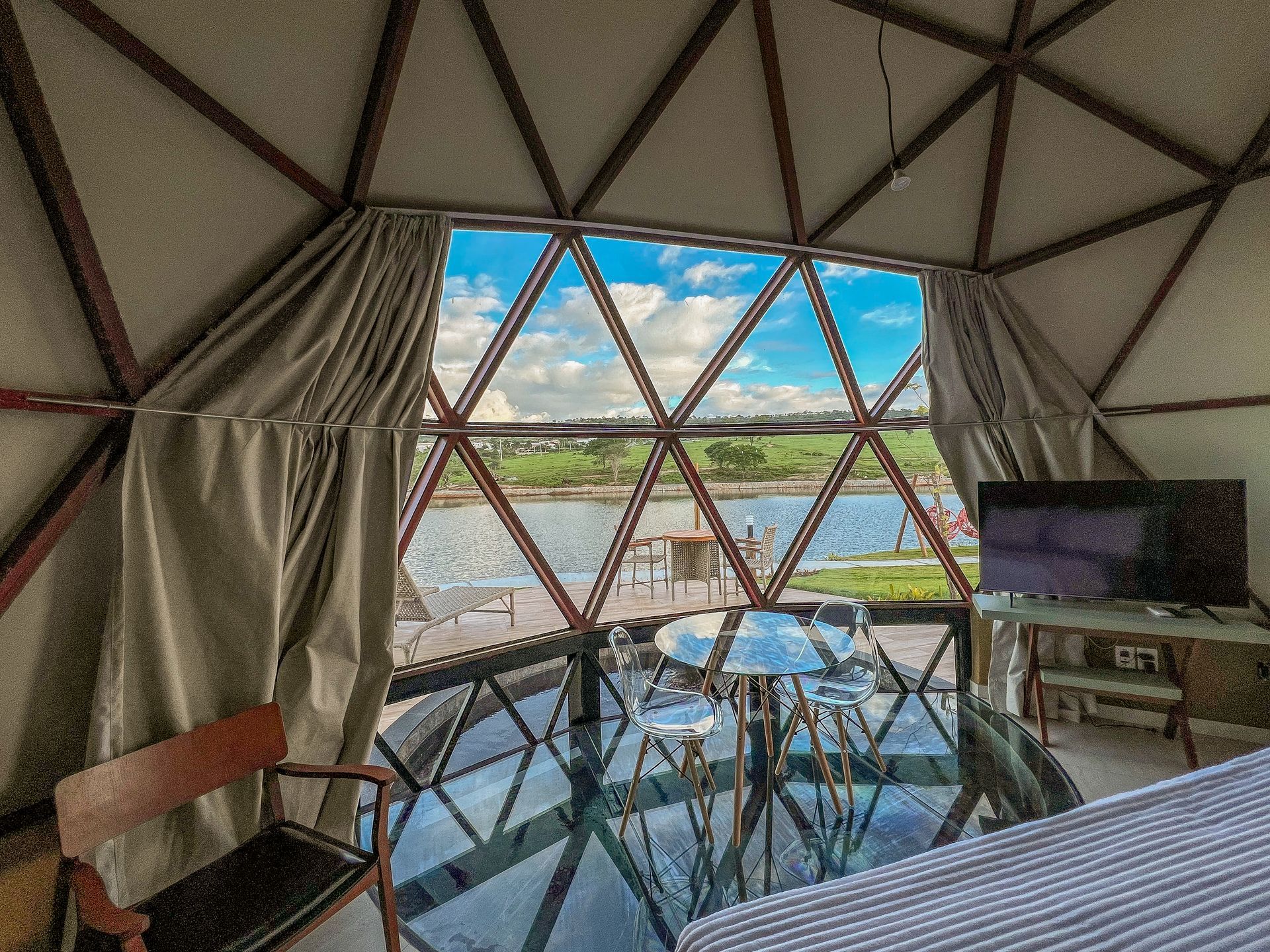 Salão em forma de cúpula geodésica com mesa de vidro, TV e vista para a cidade à beira-mar através de janelas triangulares.
