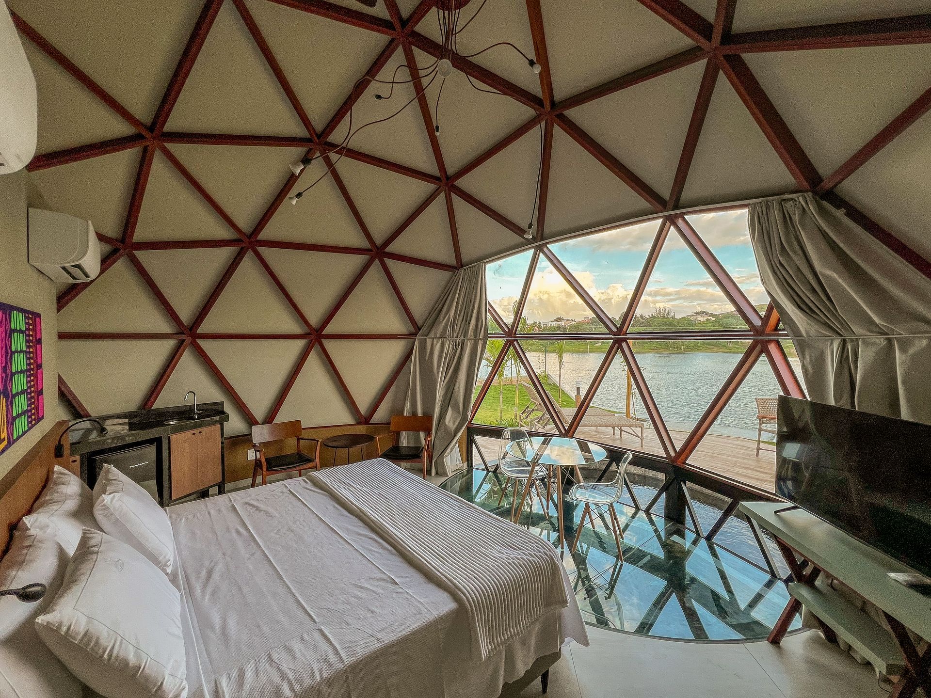 Quarto em cúpula geodésica com cama, parede de vidro e vista para o oceano ao pôr do sol.