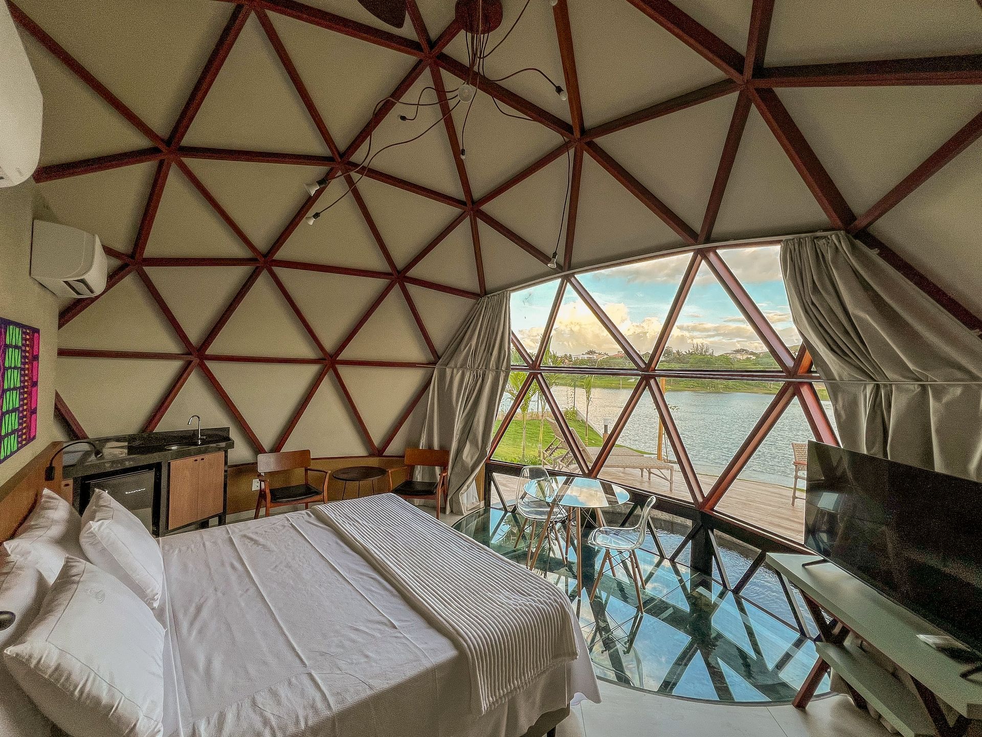 Quarto em cúpula geodésica com cama, cortinas e parede de vidro com vista para a água e as montanhas.