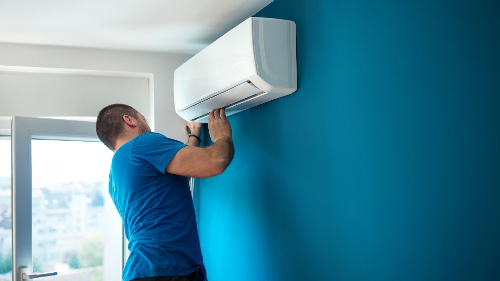 A man is installing an air conditioner on a blue wall.