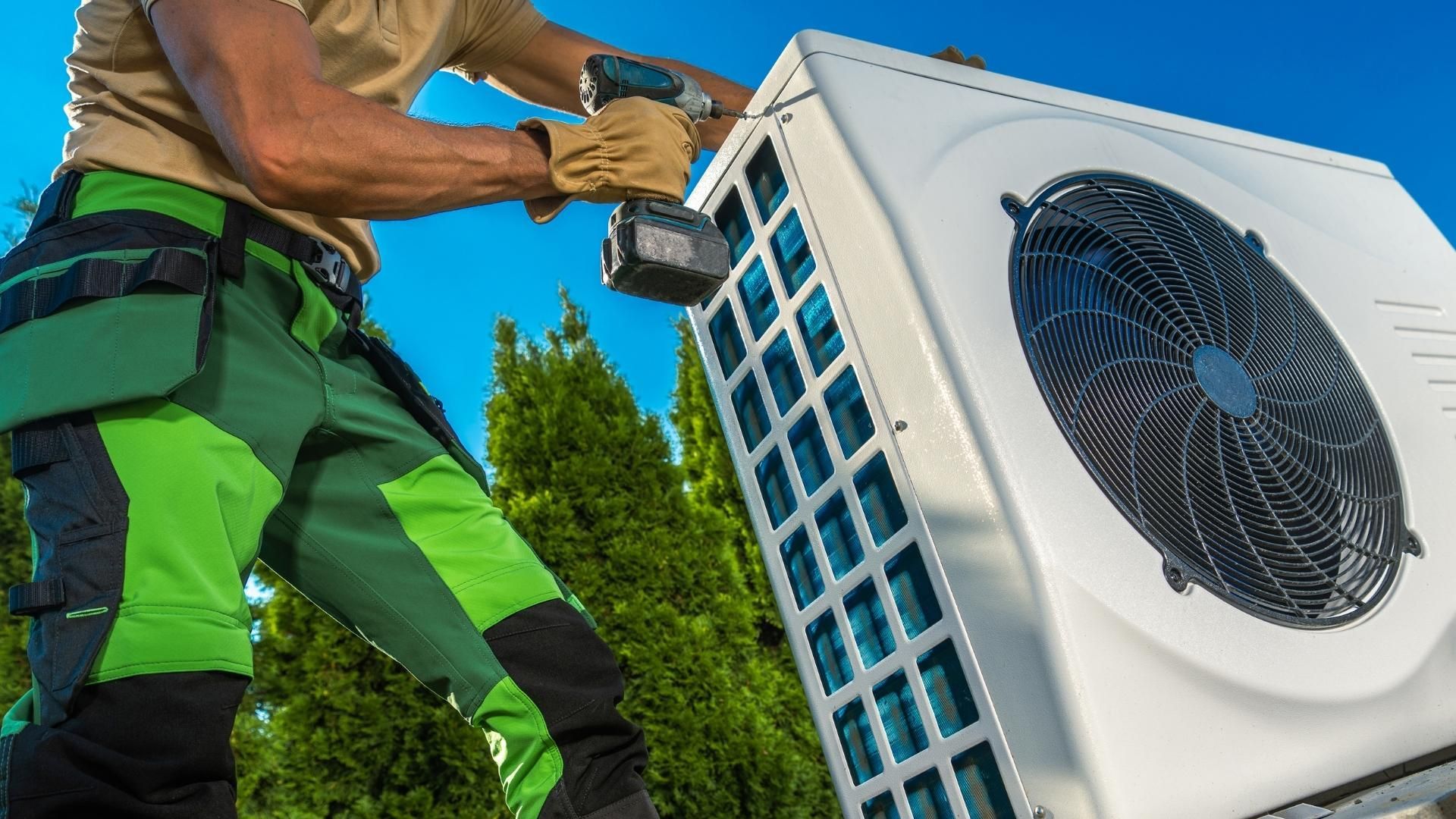 A man in green pants is working on an air conditioner with a drill.