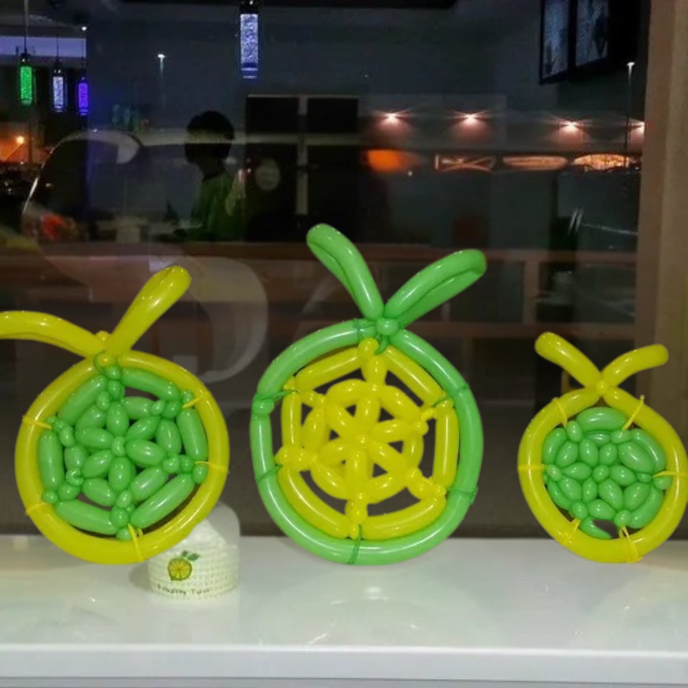 Three balloon citrus fruit decorations, yellow and green, on a ledge; a reflection in the glass behind.