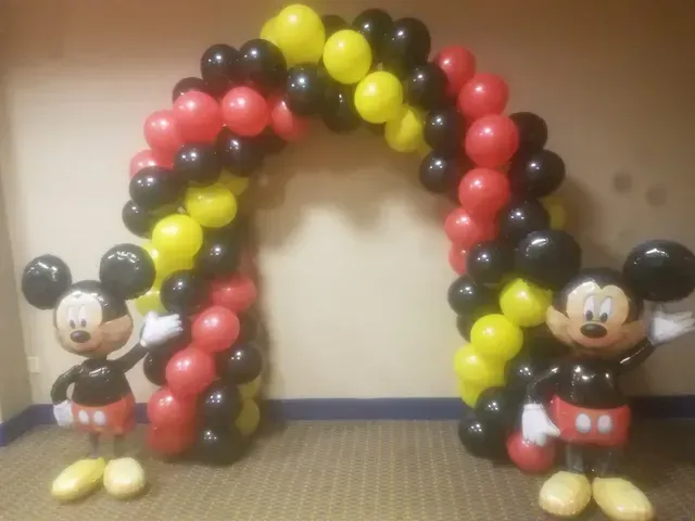 Two mickey mouse balloons are standing under a balloon arch.
