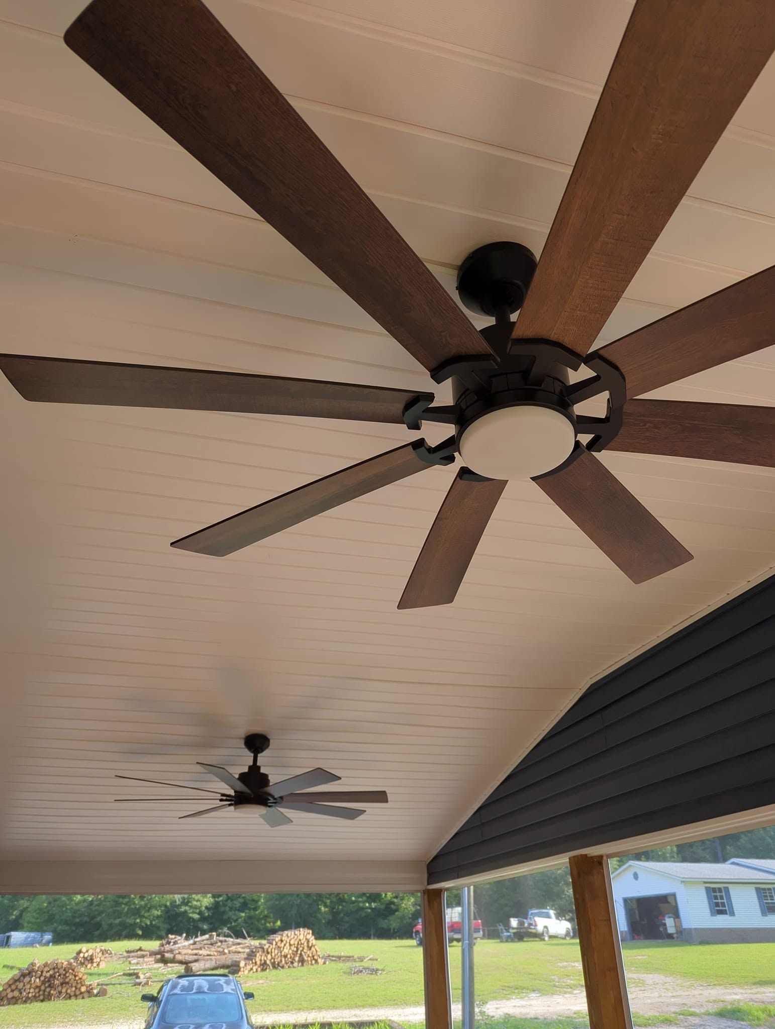 Covered porch ceiling with two ceiling fans and wooden beams, overlooking a grassy yard and house.