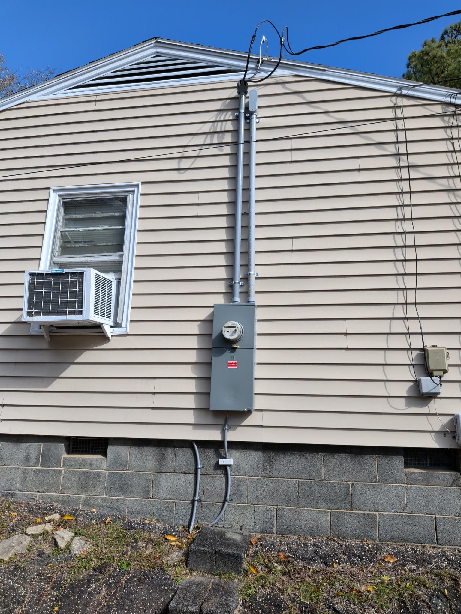House side wall with beige siding, a window air conditioner, and electrical meter boxes above a stone foundation.