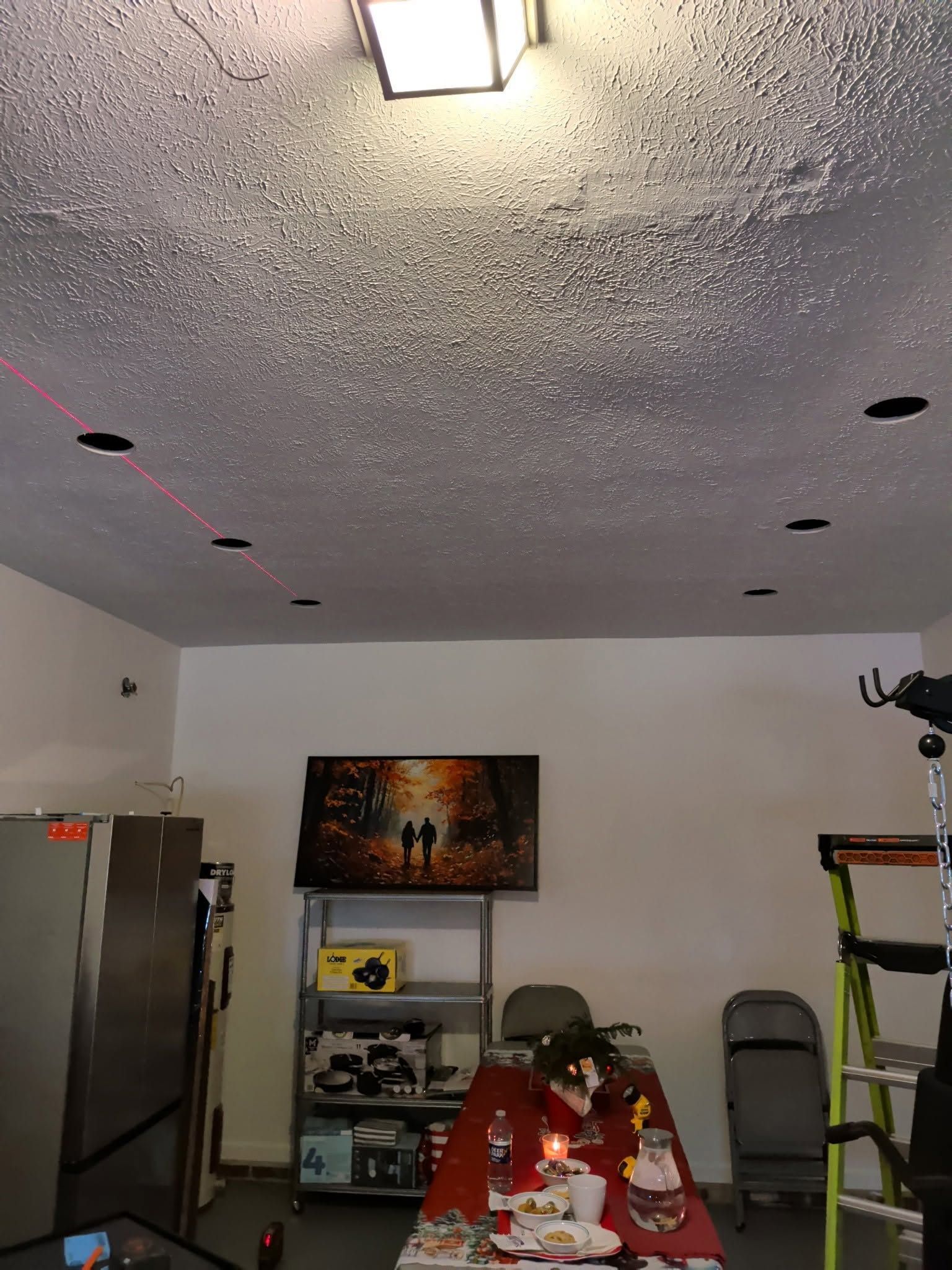 A cluttered garage with a refrigerator, shelves, chairs, and a red-covered table under a ceiling light.