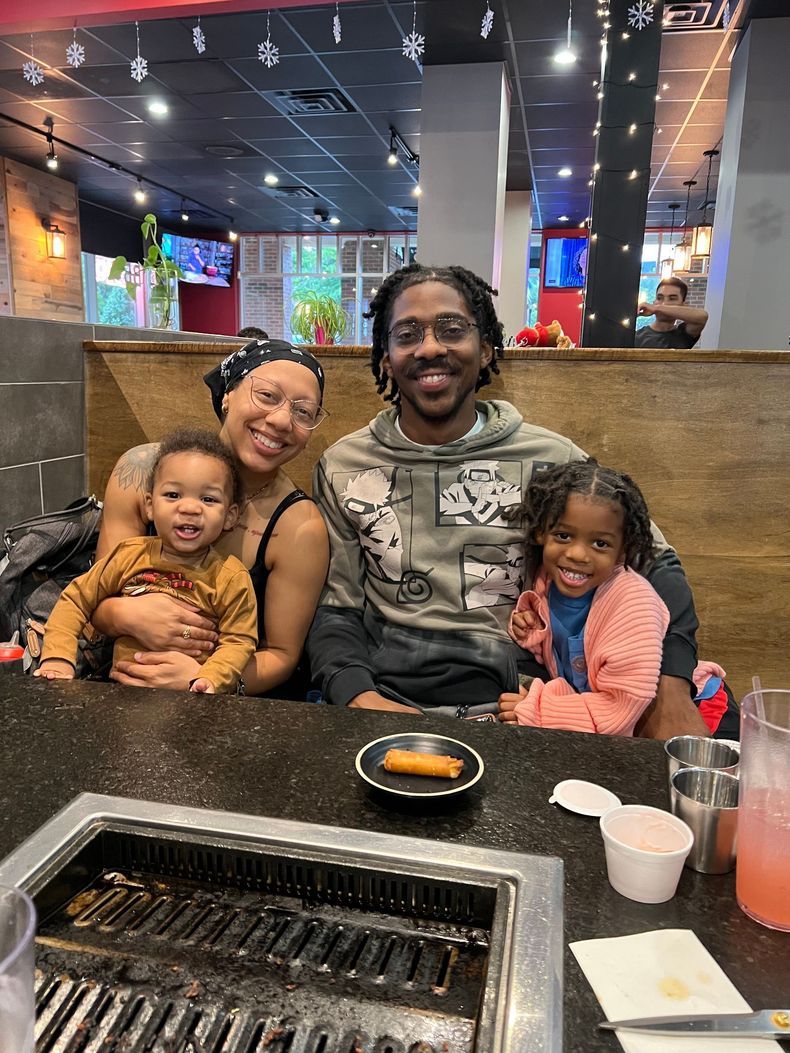 Family seated at a restaurant table, smiling with a grill in front and a bowl of food in the center